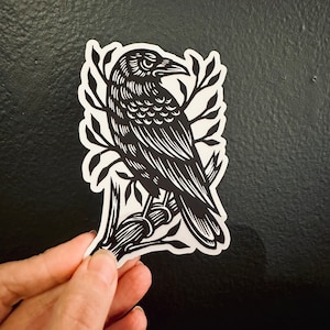 Peut inclure: Autocollant noir et blanc d'un corbeau perch&eacute; sur une branche. Le corbeau est d&eacute;taill&eacute; avec des plumes et a un bec pointu. La branche a des feuilles et est stylis&eacute;e.