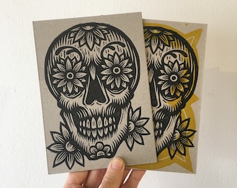 May include: Two black and white sugar skull prints on brown cardstock. The skull is decorated with floral designs. One print has a yellow background.
