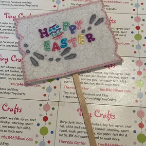 May include: A white felt applique with colorful embroidery that reads "Happy Easter" with a decorative egg and bunny rabbit designs. The applique is attached to a wooden stick.