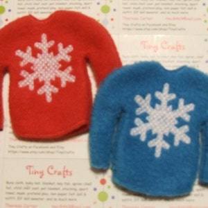 May include: Two elf-sized sweaters with snowflake designs. One sweater is red with a white snowflake, and the other is blue with a white snowflake.