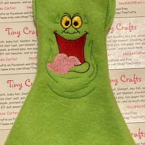 May include: Green felt doll clothing with a stitched-on cartoon face and a pink heart. The doll clothing is for a felt doll and is part of a pretend play set. The text "Tiny Crafts" is visible in the image.