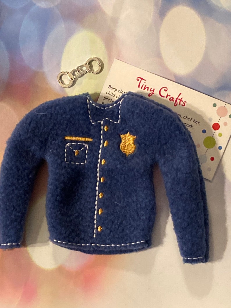 May include: A blue felt police officer costume for a doll or elf, with a gold badge and buttons. The costume is complete with a white collar and cuffs. The costume is accompanied by a pair of silver handcuffs.