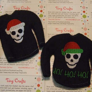 Può includere: Due maglioni di Natale in feltro nero con un teschio bianco e un cappello di Babbo Natale. Un maglione dice "HOL HO! HO!" in lettere verdi.