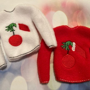 May include: Two elf-sized Christmas sweaters, one white and one red, with an embroidered image of the Grinch's hand holding a Christmas ornament.