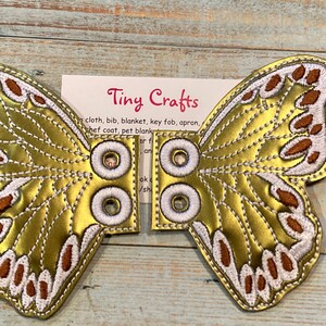 May include: Gold butterfly wings with brown and white embroidery. The wings are attached with white fabric loops and eyelets.