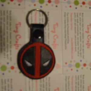 May include: A black and red key chain with a Deadpool character design. The key chain is made of fabric and has a black leather strap.