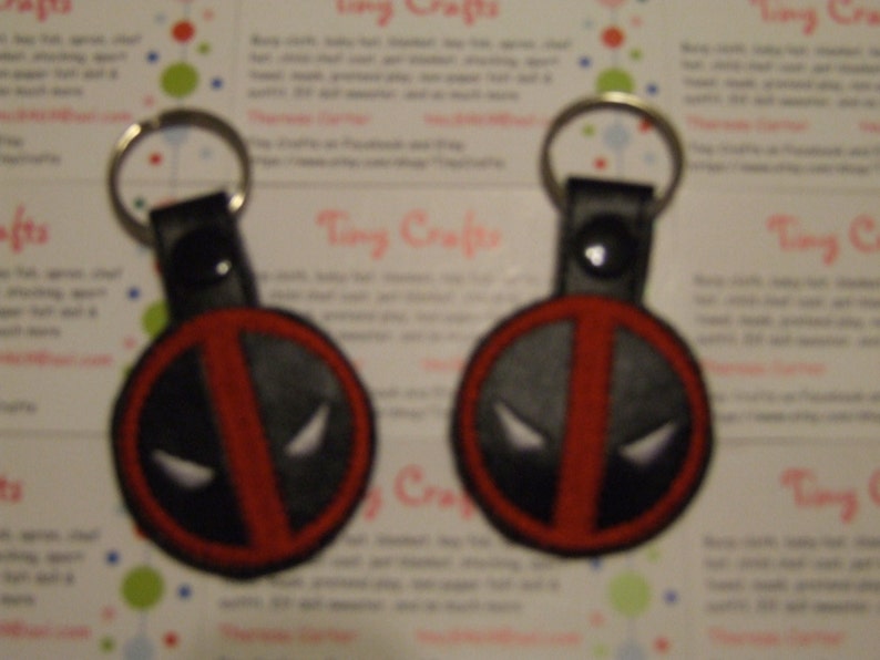 May include: Two red and black keychains with a Deadpool logo. The keychains are made of fabric and have a black leather strap.