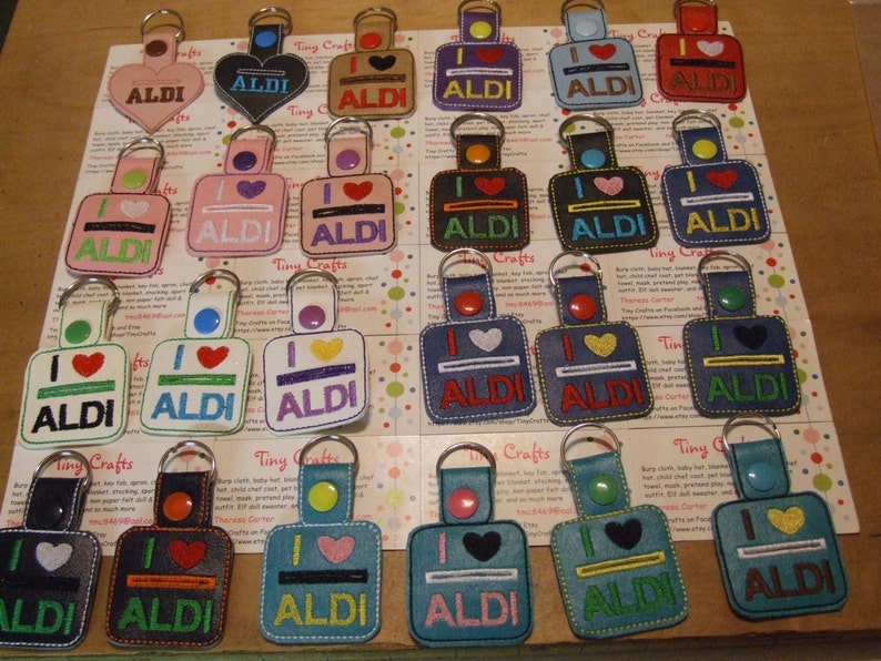 May include: A collection of 20 keychains featuring a variety of colors and designs. Each keychain has a square or heart-shaped tag with the text "I love ALDI" in white, black, or green lettering. The keychains are made of leather and have a metal ring.