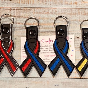 May include: Four keychains with black leather straps and embroidered ribbon designs. The ribbons are red and blue, blue and blue, blue and yellow, and red and black.
