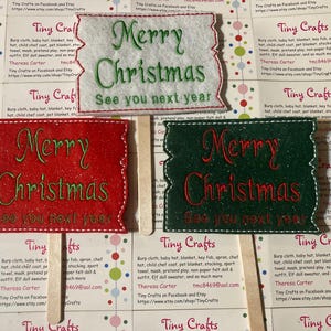 May include: Three felt Christmas signs with the text "Merry Christmas See you next year" in red and green lettering on a white, red, and green background. The signs are attached to wooden sticks.