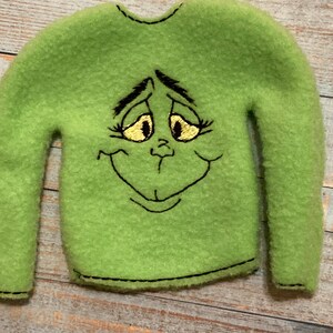 May include: Green fleece elf-sized sweater with an embroidered Grinch face. The Grinch has black eyebrows, eyelashes, and a mouth, and yellow eyes.
