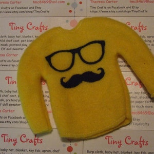 Puede incluir: Un suéter de fieltro amarillo con diseño de gafas y bigote negros. El suéter es un artículo de artesanía para muñecas. El fondo incluye tarjetas de visita con el texto "Tiny Crafts" e información de contacto.