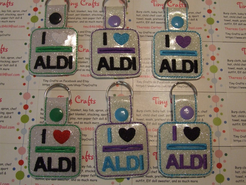 May include: A set of eight keychains with a white background and glitter. Each keychain features a different colored heart and a line with the text "I love ALDI" in black letters.