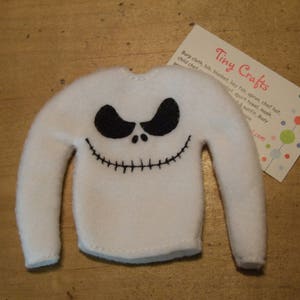Puede incluir: Suéter de fieltro blanco de tamaño de duende con una cara de Jack Skellington bordada en negro. El suéter tiene mangas largas y cuello redondo.