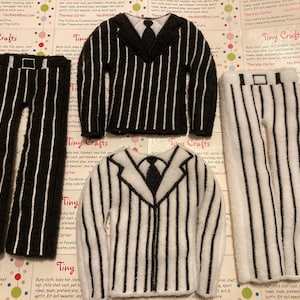 Op de afbeelding: Een set van vier vilten poppenkleding in een zwart-wit gestreept patroon. De set bevat een jas, een broek en twee shirts. De kleding is gemaakt van vilt en is perfect voor een poppenhuis of voor rollenspel.