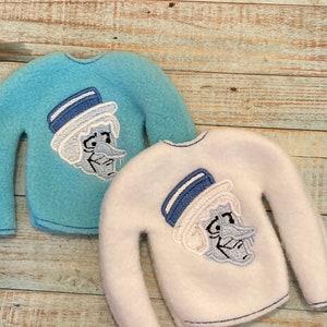 May include: Two elf-sized sweaters featuring a blue and white embroidered cartoon character wearing a hat. The sweater on the left is light blue and the sweater on the right is white.