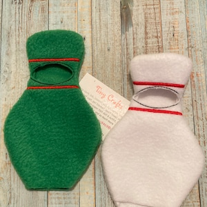 Pode incluir: Dois chapéus de boneca de feltro, um verde e outro branco, com acabamento vermelho. Os chapéus são em forma de garrafa e têm um buraco para a cabeça de uma boneca. Os chapéus estão sobre um fundo de madeira. Tiny Crafts está escrito em um cartão na imagem.