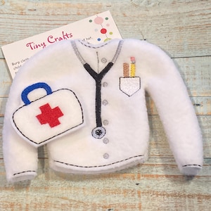 May include: White felt doctor costume for a doll or elf. The costume features a stethoscope, a pocket with pencils, and a small red cross first aid kit.