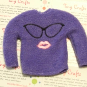 May include: A purple felt sweater with black glasses and pink lips embroidered on the front. The sweater is designed for a doll and has long sleeves. The image also includes text from "Tiny Crafts".
