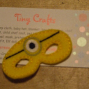 May include: A yellow felt eye mask with a black felt strap and a blue eye detail. The mask is shaped like a minion from the Despicable Me movies. The mask is on a white card with the text "Tiny Crafts" in pink.