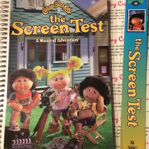 May include: A Cabbage Patch Kids book titled "The Screen Test: A Musical Adventure" featuring three Cabbage Patch Kids dolls. The dolls are dressed in colorful outfits and are holding props, such as a camera and a clapperboard. The book cover is a bright blue and yellow color scheme.