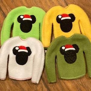 May include: Four elf-sized sweaters with a Mickey Mouse design. The sweaters are green, yellow, white, and dark green.