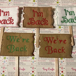 Op de afbeelding: Vier viltborden met gegolfde randen, twee bruin, een wit en een beige. De borden hebben de tekst "I'm Back" en "We're Back" geborduurd in rood en groen garen.