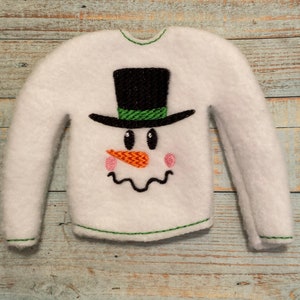 May include: White long-sleeved sweater with a green embroidered snowman design. The snowman has a black top hat with a green band, orange carrot nose, and black eyes.