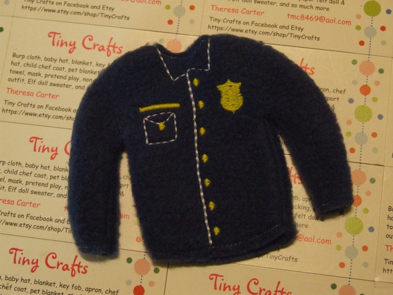May include: A blue felt police officer's jacket with yellow stitching and a gold badge. The jacket is a felt applique for a doll or craft project. Tiny Crafts on Facebook and Etsy, tmc8469@aol.com
