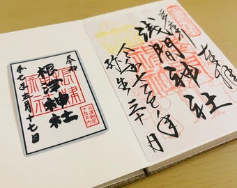 May include: A white notebook open to two pages with Japanese calligraphy and red stamps. One page features a rectangular design with Japanese text and a red stamp. The other page has more calligraphy and a red stamp.