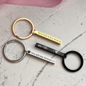 Könnte beinhalten: Zwei goldene und schwarze Schlüsselanhänger mit gravierten Balken. Der goldene Balken trägt die Aufschrift "40°41 21.15 N, 74°2 39.93 W". Der schwarze Balken trägt die Aufschrift "You are my forever" mit einem Schlüssel- und Herzsymbol.