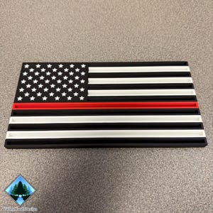 May include: A rectangular, 3D-printed American flag with a red line. The flag features a field of white stars on a blue background, black and white stripes, and a prominent red stripe. The design is a patriotic symbol.