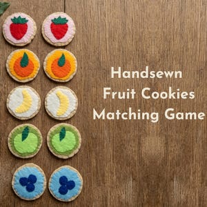 May include: A handsewn fruit cookies matching game featuring ten circular felt pieces arranged in two columns. Each piece displays a different fruit design including strawberry, orange, banana, apple, and blueberry. The text reads Handsewn Fruit Cookies Matching Game.