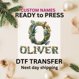 May include: A white background with the text "CUSTOM NAMES READY to PRESS" in pink and black. A white square with a green and brown letter "O" surrounded by leaves and a dinosaur. The word "OLIVER" is written below the letter "O" in green and brown letters. The text "DTF TRANSFER" is written in black below the square. The text "Next day shipping" is written in black below the text "DTF TRANSFER".