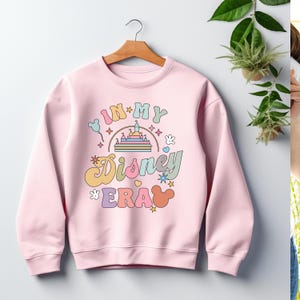 May include: A pale pink sweatshirt featuring the phrase "IN MY Disney ERA" in pastel hues. The design incorporates a castle, Mickey Mouse ears, stars, and other playful motifs. The sweatshirt is hung on a wooden hanger.