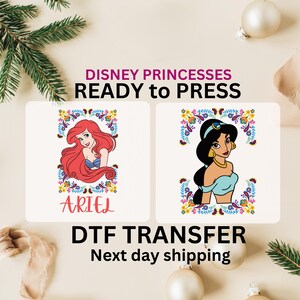 May include: Two printable iron-on transfers featuring Disney princesses Ariel and Jasmine. The images are surrounded by colorful floral borders. The text "DISNEY PRINCESSES READY to PRESS DTF TRANSFER Next day shipping" is displayed below the images.