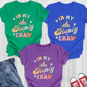 May include: Three t-shirts in green, blue, and purple, each with the text "IN MY Disney ERA" in a retro font. The design includes a castle, Mickey Mouse ears, and stars. The shirts are folded on a light wood surface.
