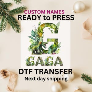 May include: A DTF transfer featuring a watercolor design with the name "GAGA" and a large letter "G" decorated with green foliage and a dinosaur. The text "CUSTOM NAMES READY to PRESS" is at the top.