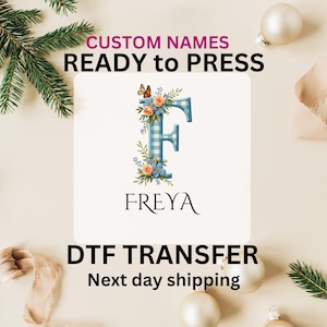 May include: A white square with a blue and white checkered letter F surrounded by flowers and butterflies. The name "FREYA" is written below the letter. The text "CUSTOM NAMES READY to PRESS DTF TRANSFER Next day shipping" is written above and below the image.