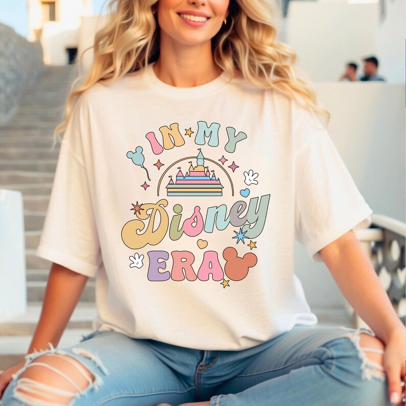 May include: White t-shirt featuring the phrase "IN MY Disney ERA" in a retro, colourful font. The design includes a castle, Mickey Mouse ears, stars, and other playful elements. A casual, everyday wear item.