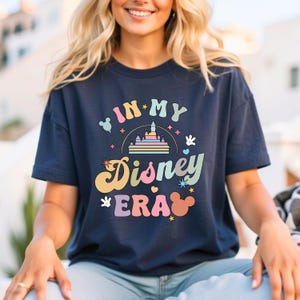 May include: Navy blue t-shirt featuring the text "IN MY Disney ERA" in a retro font with a castle, Mickey Mouse ears, and other Disney-themed graphics. The shirt is a casual style, perfect for Disney fans. The design is colourful.