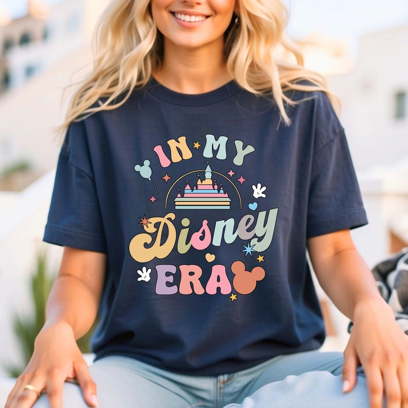 May include: Navy blue t-shirt featuring the text "IN MY Disney ERA" in a retro font with a castle, Mickey Mouse ears, and other Disney-themed graphics. The shirt is a casual style, perfect for Disney fans. The design is colourful.