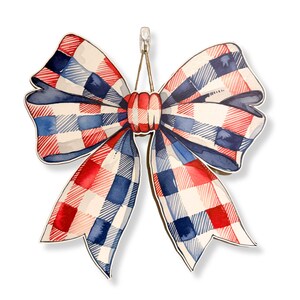 May include: A decorative bow with a patriotic theme. The bow features a red, white, and blue plaid pattern. It is hanging from a metal hook. The bow is made of a sturdy material and has a watercolor effect.