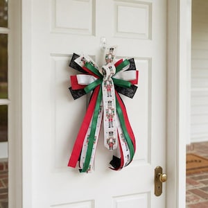 May include: A decorative bow made of layered ribbons in red, green, white, and black. The central ribbon features a repeating pattern of nutcracker figures. The bow includes solid red, solid green, and black ribbons with a subtle swirl pattern.