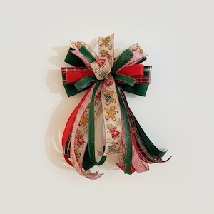 May include: A decorative Christmas bow featuring a mix of ribbons in red, green, and beige. The bow includes a plaid pattern, solid colors, and a gingerbread man design. The bow is suitable for holiday decorations.