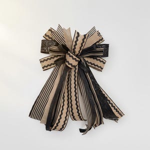 May include: A decorative bow featuring a mix of black and tan ribbons. The bow has a variety of patterns, including stripes, zigzags, and embossed floral designs. The ribbons are arranged to create a full, layered look, suitable for various decorative purposes.