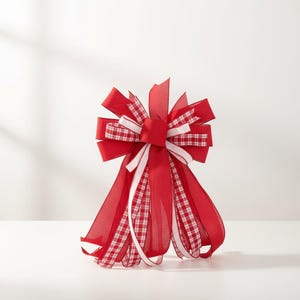 May include: A large, decorative bow featuring a mix of red, white, and plaid ribbons. The bow has multiple layers and streamers, perfect for holiday or gift wrapping. The bow is made of fabric and has a festive appearance.