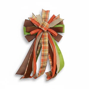 May include: A decorative bow featuring a mix of ribbons in brown, green, orange, and tan. The bow has a plaid center and long, flowing ribbons. Perfect for fall or seasonal decor.