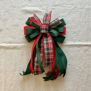 May include: A decorative Christmas bow featuring a plaid pattern in red, green, and white. The bow has multiple layers of ribbon, with long, flowing tails in solid red and green. The bow is designed for holiday decorations.