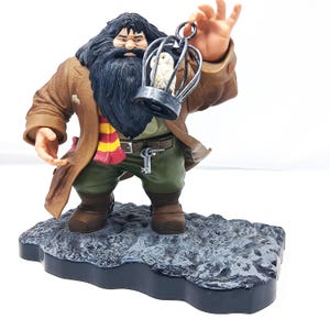 May include: A figurine of a character with a long black beard, wearing a brown coat, green pants, and a scarf with red and yellow stripes. The character is holding a cage with a white owl inside. The figurine is standing on a gray, textured base.